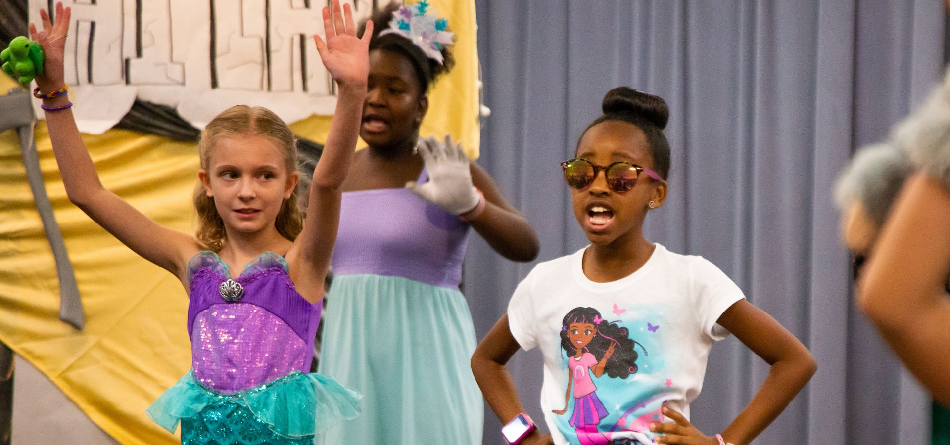 Three girls perform on stage; one wears a mermaid costume, another in a purple dress stands behind, and the third wears sunglasses and a T-shirt with a cartoon character.
