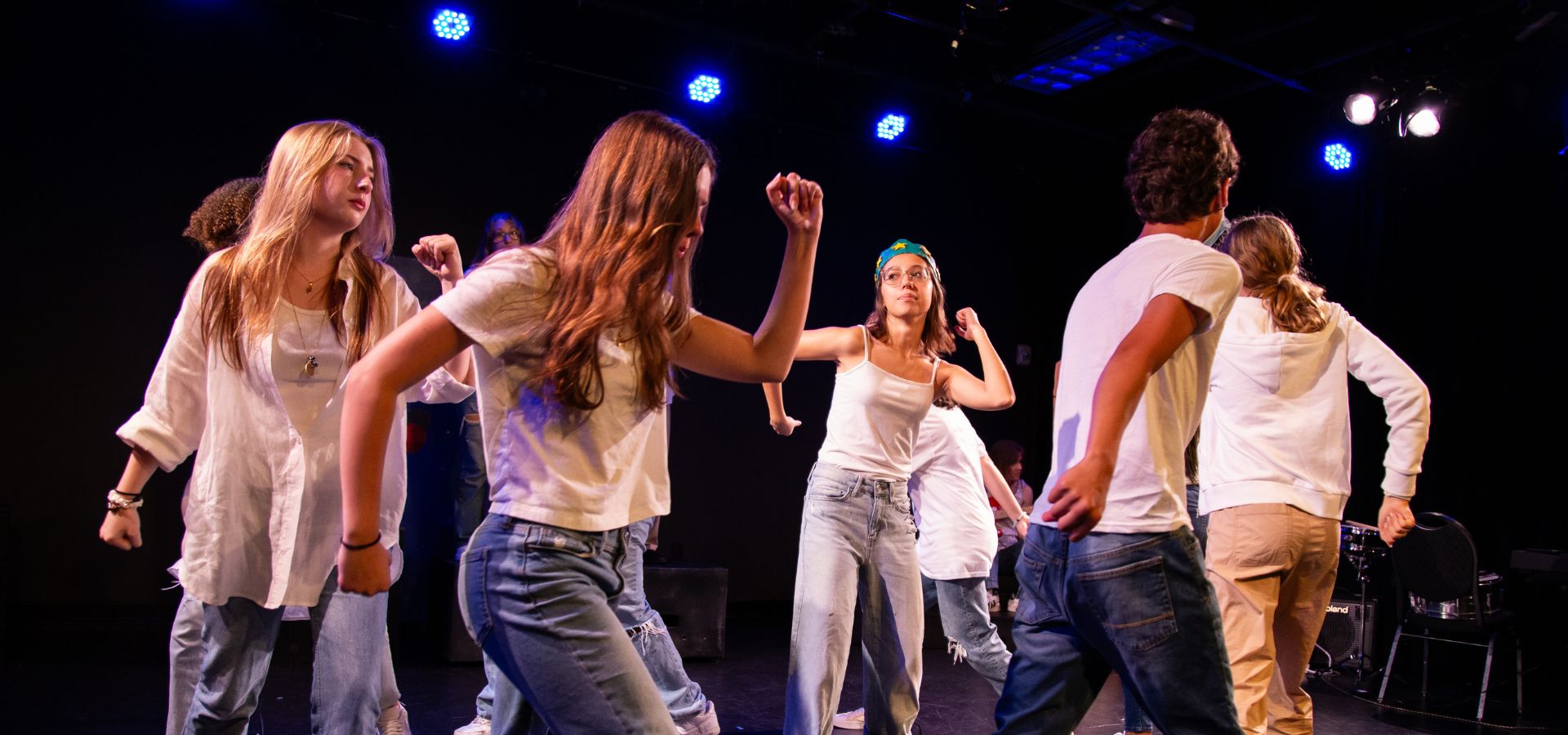 A group of young people in casual clothes perform a choreographed dance on a stage under blue and white lights.