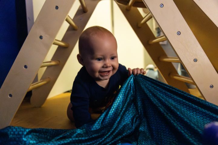 A baby crawls under a wooden climbing frame, smiling and holding a blue textured fabric.