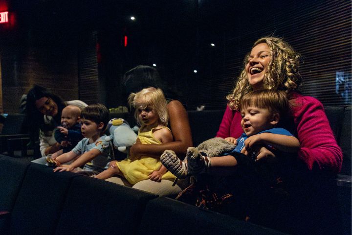 Adults and young children sit together in a dimly lit room, some holding stuffed animals, while one woman laughs and holds a smiling toddler on her lap.
