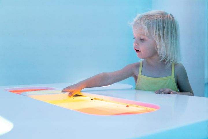 Young child in a yellow tank top touches a glowing yellow-lit surface on a white table in a bright blue room.