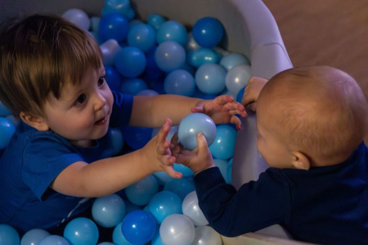 Two young children play together in a ball pit filled with blue and light blue plastic balls, reaching toward each other.