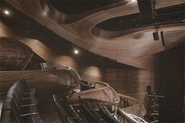 Modern theater interior with black seating and intricate, curved wooden architectural details on the walls and ceiling, with stage lighting equipment visible to the right.