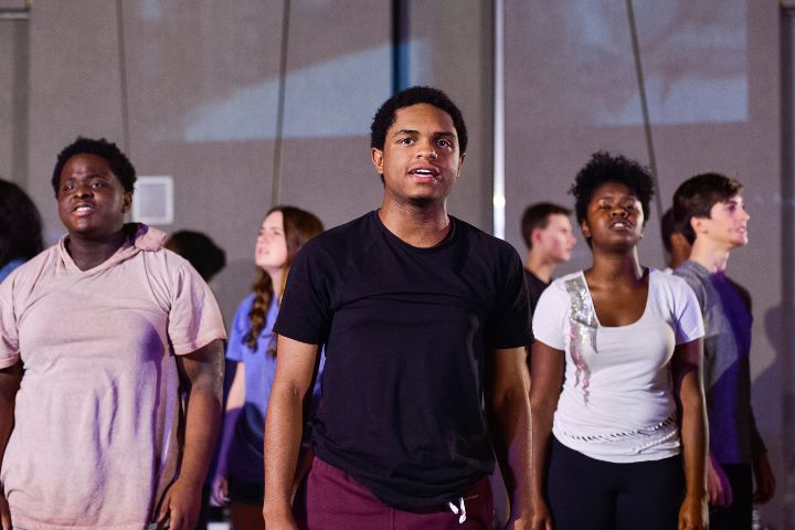 A group of young people stand in rows, facing forward, appearing to participate in a performance or rehearsal.
