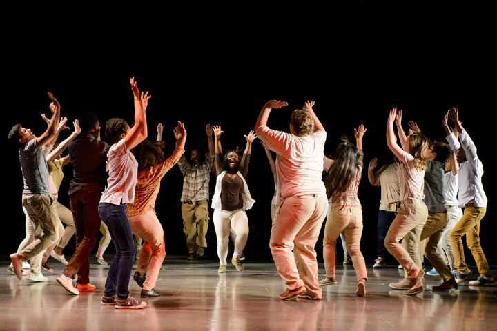 A group of people stands in a circle, raising their arms and moving energetically on a stage with a dark background.
