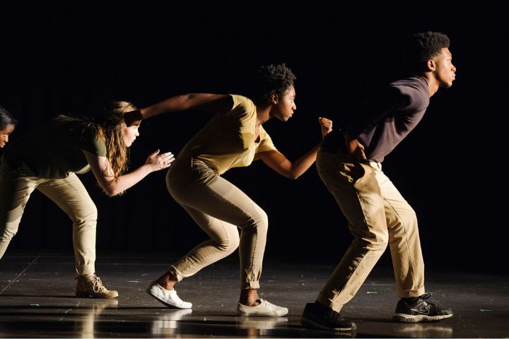Three people on stage, dressed in neutral clothing, strike dynamic poses as if in motion, leaning forward with arms extended, against a dark background.