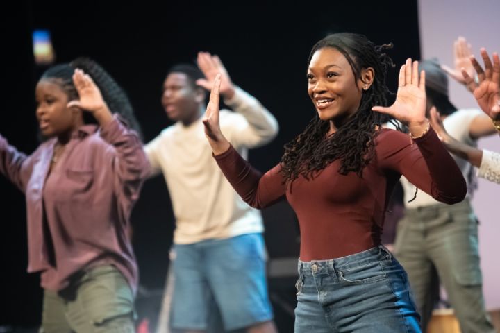 A group of young people on stage perform a dance routine, raising their hands by their heads; a smiling woman in a maroon top is in the foreground.