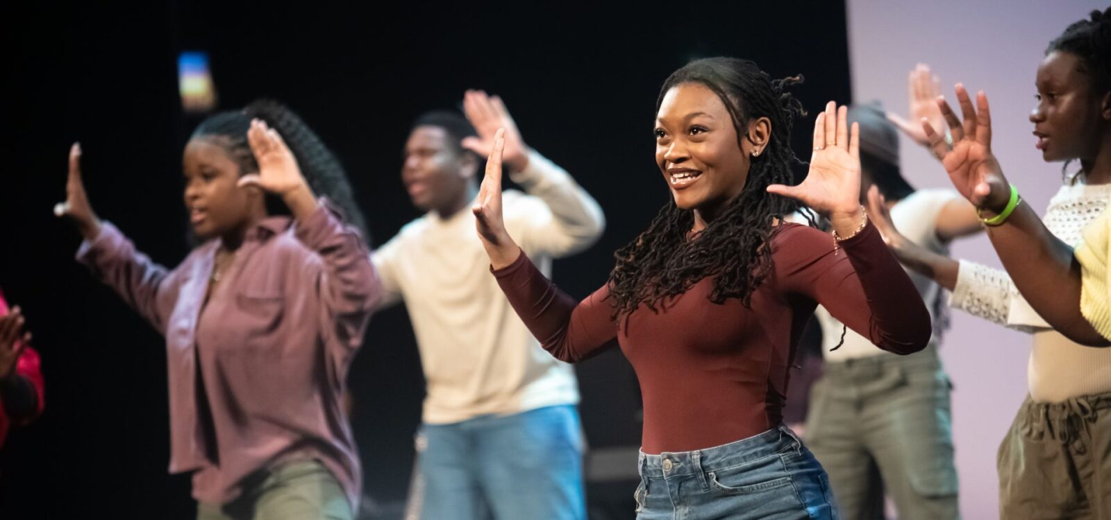 A group of people stand in a row on stage, raising their hands with open palms, appearing to rehearse or perform a dance routine.