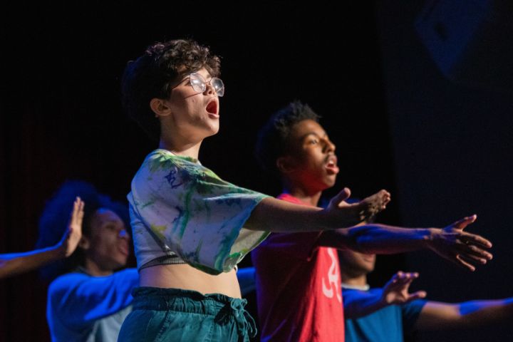 A group of young people with expressive faces stand in a row on stage, extending their arms forward as they perform.