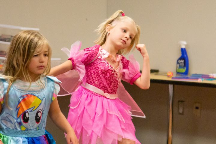 Two young girls in colorful costumes, one dressed as a fairy with wings and another in a blue dress, stand indoors near a table with cleaning supplies in the background.