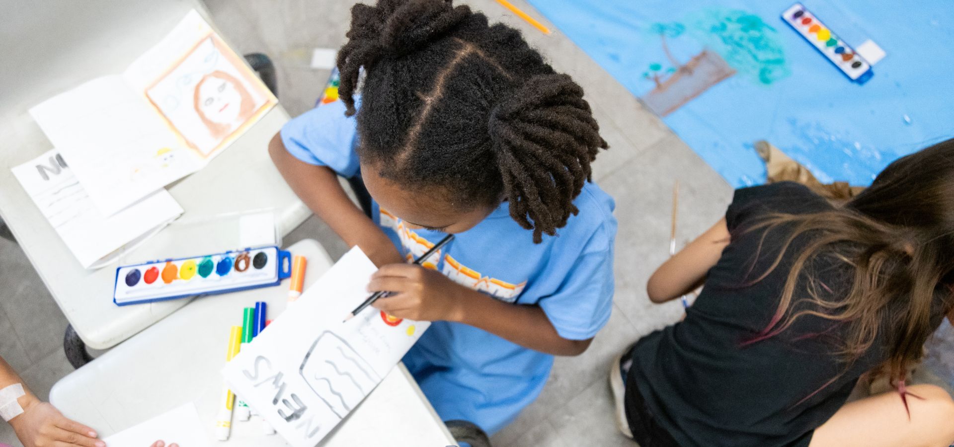 A child paints on paper at a table with art supplies, while another child sits nearby on the floor with a blue mat and art materials.