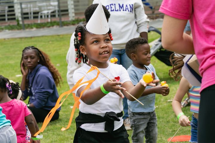 Farmers Market Young girl wearing a party hat and holding ribbon sticks smiles at an adult during an outdoor event with other children playing in the background.
