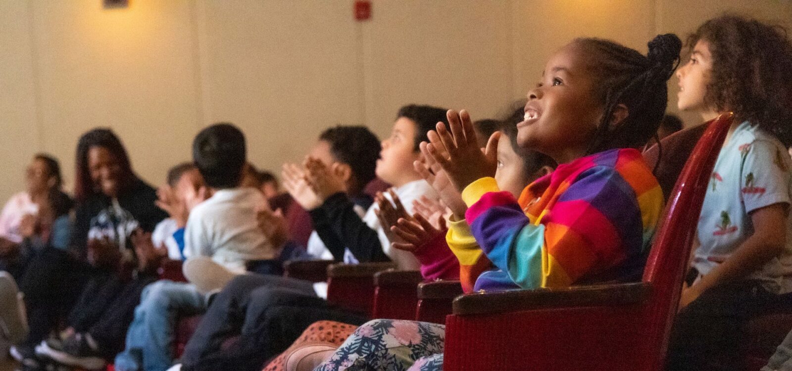 Children sit in theater seats, clapping and watching something off-screen with interest. The focus is on a girl in a colorful jacket smiling and applauding.