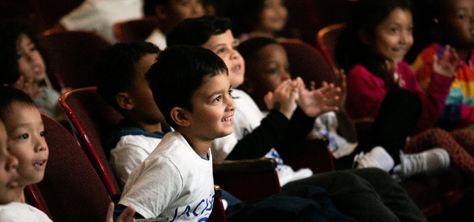 A group of children sit in theater seats, smiling and clapping while watching a performance.