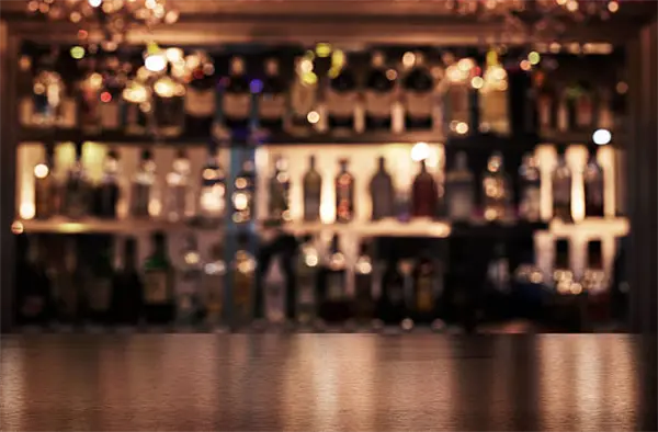 A wooden bar counter with a blurred background of various bottles and glasses on shelves, softly lit by warm lighting.