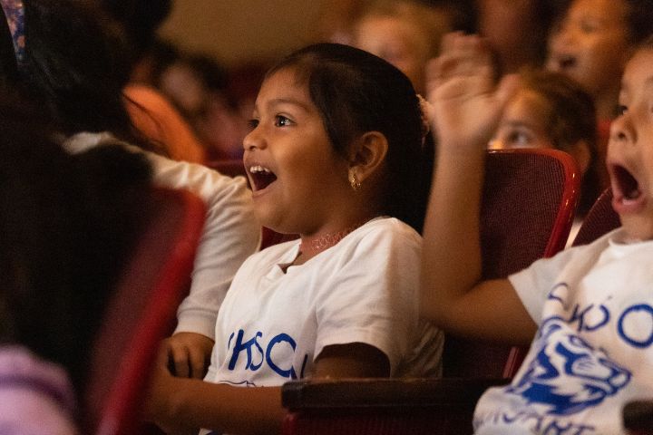 Children wearing white shirts sit in red theater seats, smiling and reacting with excitement while watching a performance or show.