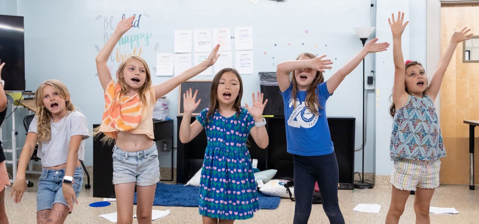 Five young girls standing indoors, singing or performing with their arms raised and expressive faces in a classroom setting.