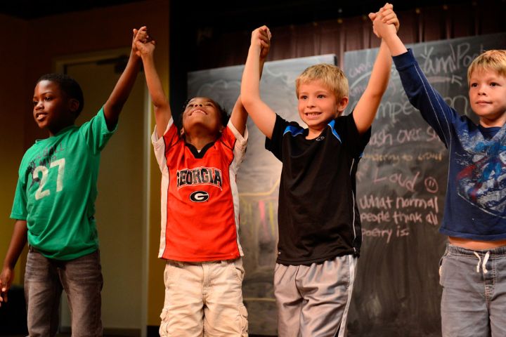 Four young boys stand on a stage holding hands and raising their arms in celebration, with a chalkboard in the background.