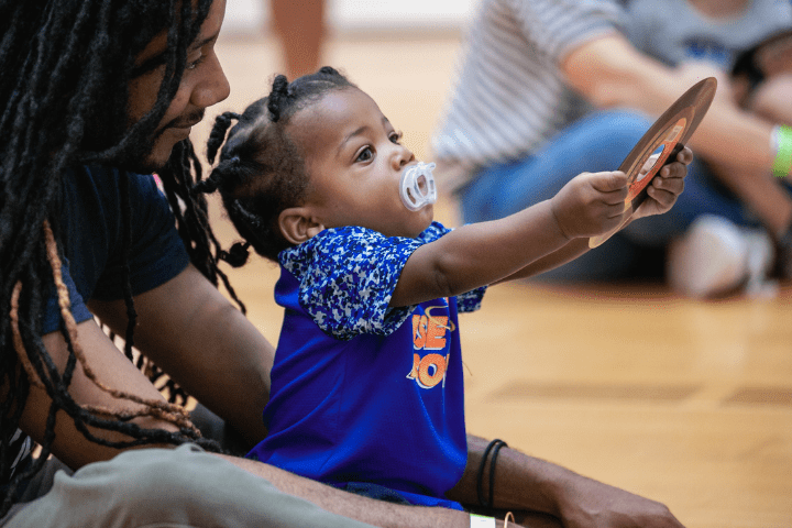 A baby with a pacifier sits on the floor holding up a round object, with an adult sitting close beside them.