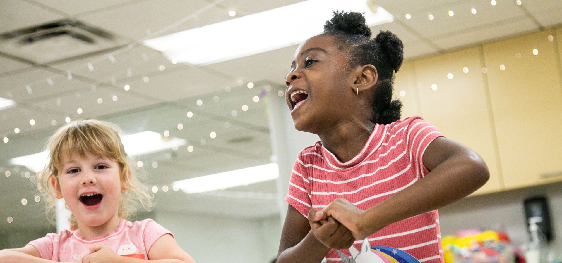 Two young girls laugh and play together indoors, holding onto a colorful object, with string lights and cabinets in the background.