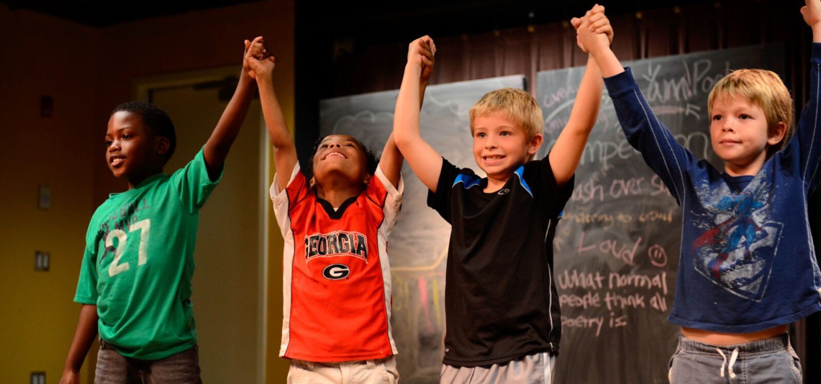 Four young boys stand on stage holding hands and raising their arms in celebration, with a chalkboard in the background.