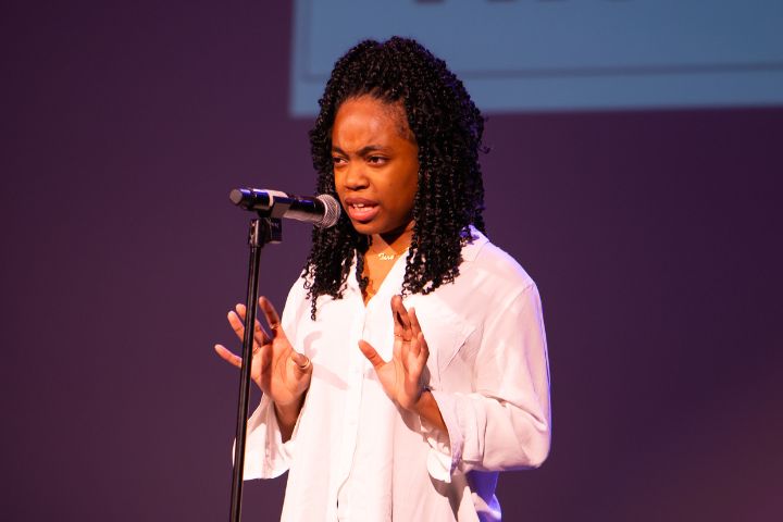 A woman in a white shirt speaks passionately at a microphone on stage, gesturing with both hands against a purple background.