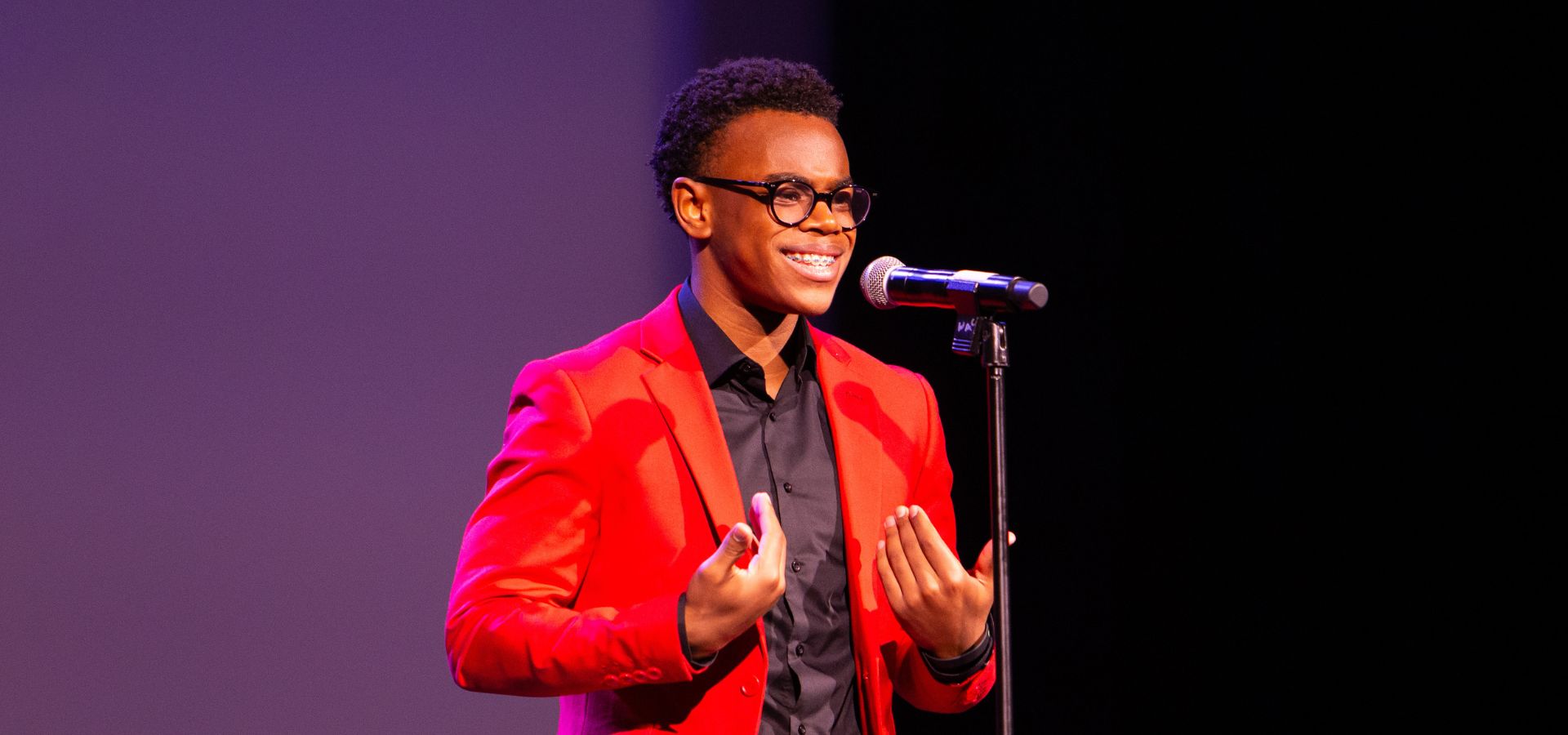 A person wearing a red jacket and black shirt speaks animatedly into a microphone on stage against a dark and purple backdrop.