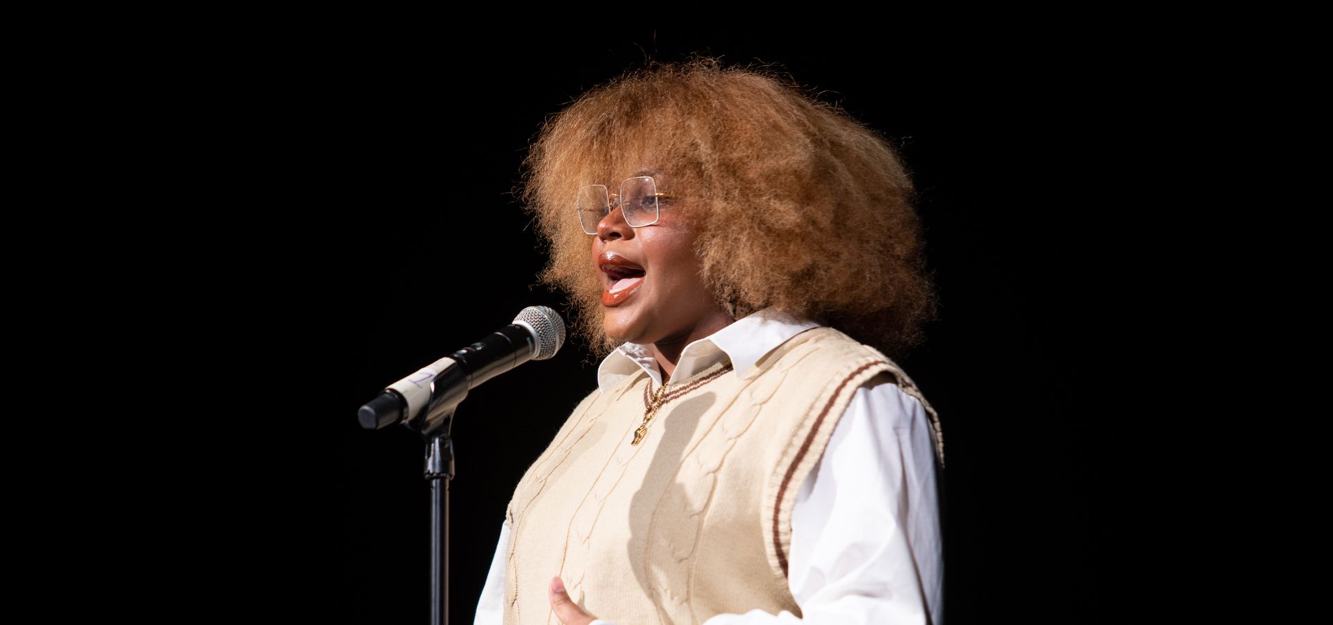 A person with curly hair and glasses sings or speaks into a microphone while wearing a light-colored sweater vest over a white shirt against a black background.