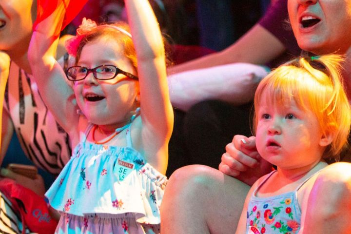 Two young children sit closely together in a crowd, one wearing glasses and raising their arms, both appearing engaged and excited by something off-camera.