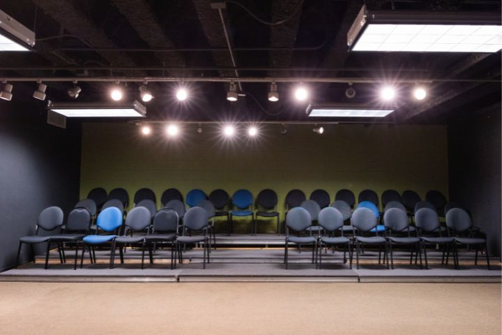 Rows of empty black and a few blue chairs are arranged on tiered platforms in a small room with ceiling lights and exposed pipes.