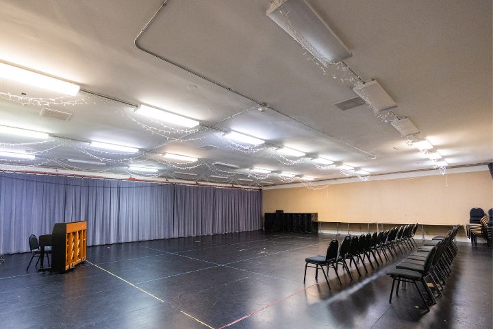 Large multipurpose room with black flooring, rows of black chairs, a piano, gray curtains along one wall, and string lights on the ceiling.