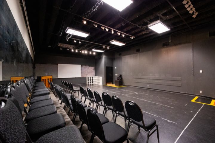 A small black box theater with rows of empty chairs facing a stage area, black walls, overhead lighting, and a piano in the corner.