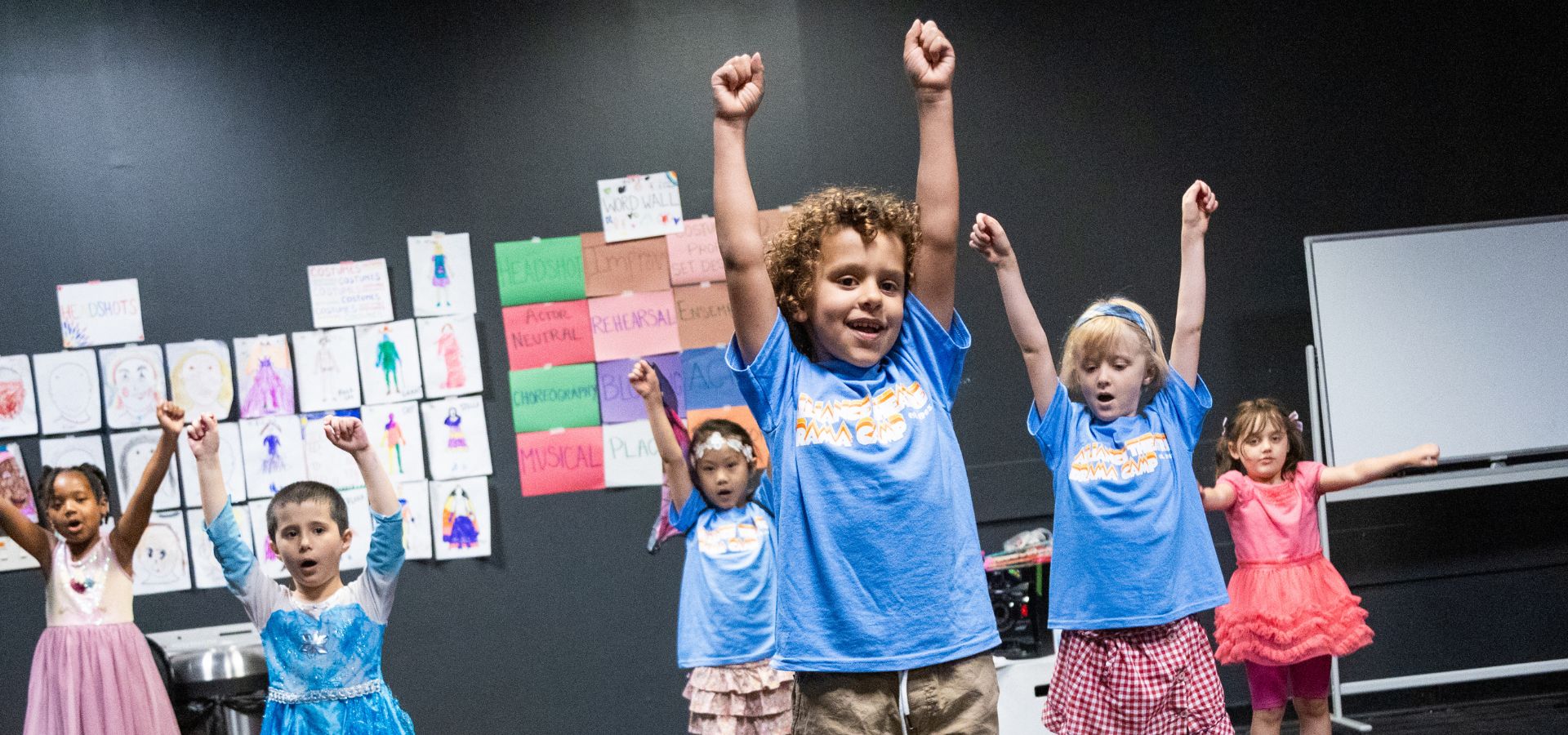 Children stand with arms raised in a classroom, some wearing blue t-shirts and others in costumes, with drawings and posters on the wall behind them.