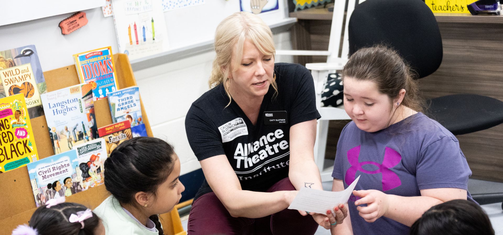 A woman sits with three young students in a classroom, helping them read a paper; children's books are visible on a shelf nearby.