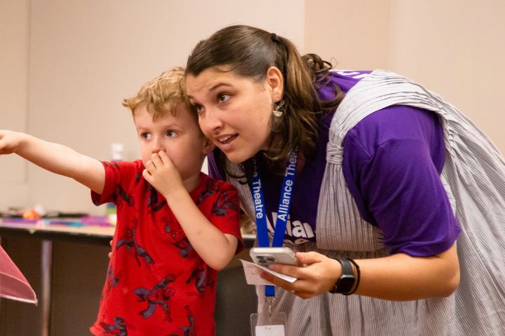 A young boy in a red shirt points at something while standing next to an adult woman with a lanyard, who is looking in the same direction.