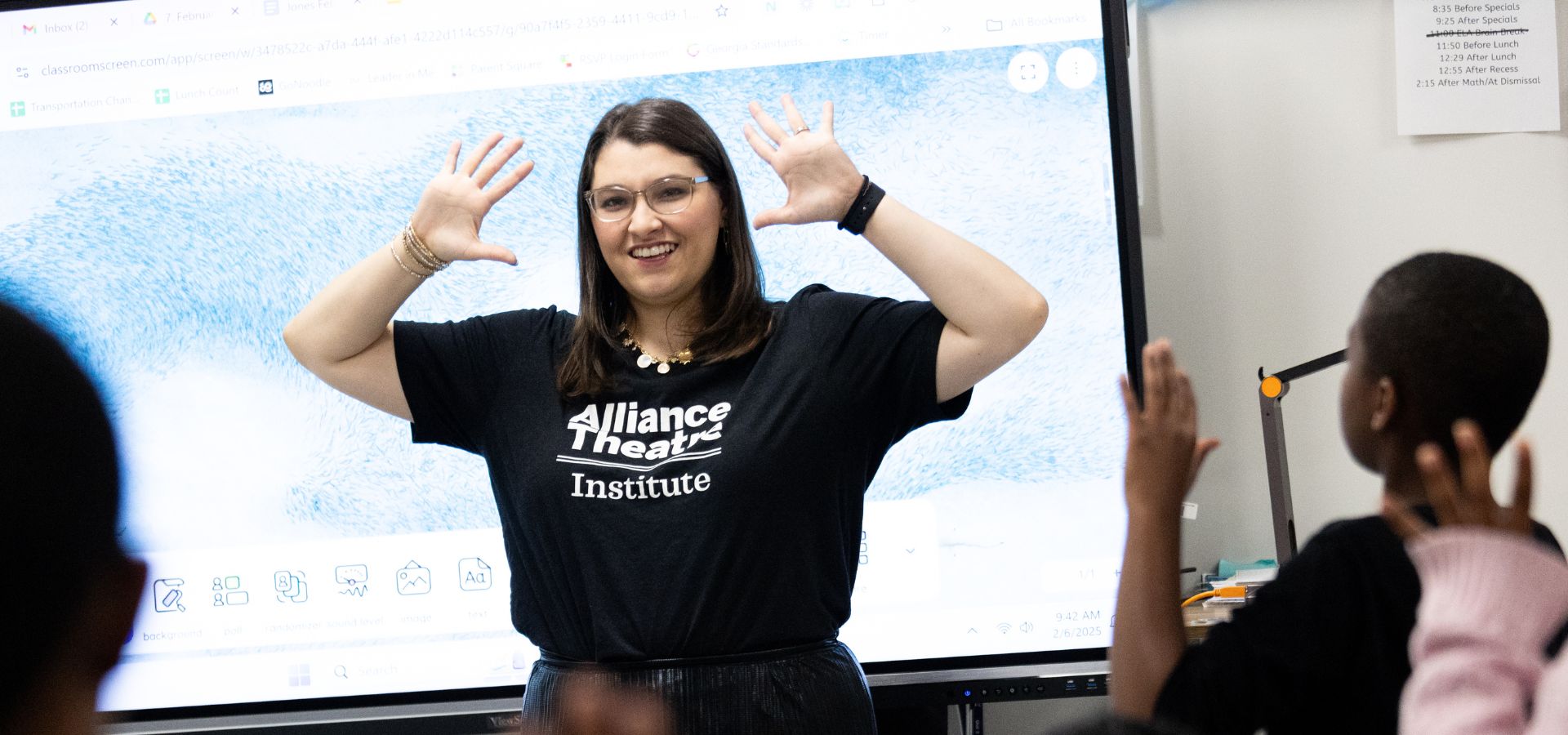 A woman wearing an "Alliance Theatre Institute" shirt stands in front of a screen, raising her hands, while children raise their hands toward her in a classroom setting.