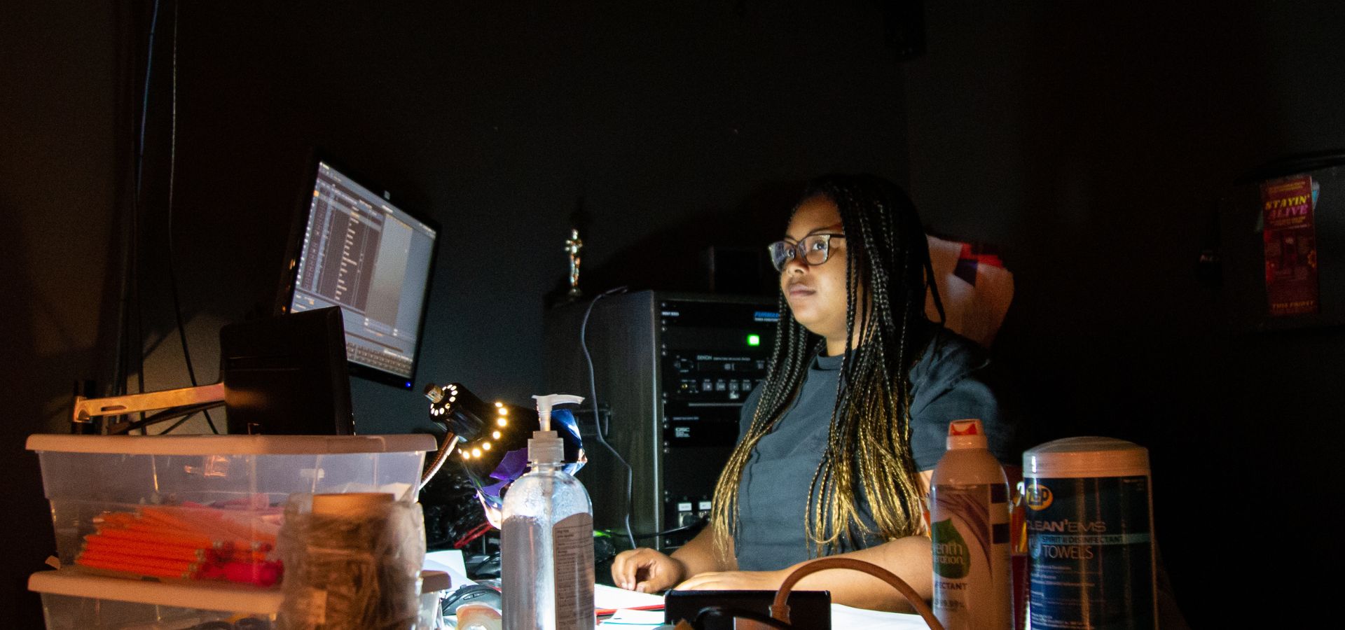 A woman with braids sits at a desk with computer monitors, electronic equipment, and office supplies in a dimly lit room.