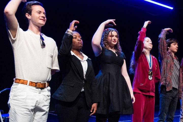 Five people stand in a row on stage, each raising one arm with a bent hand gesture under blue lighting.