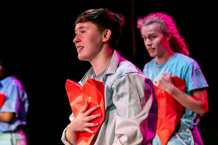 Two people on stage hold red props to their chests while performing; both wear casual costumes under stage lighting.