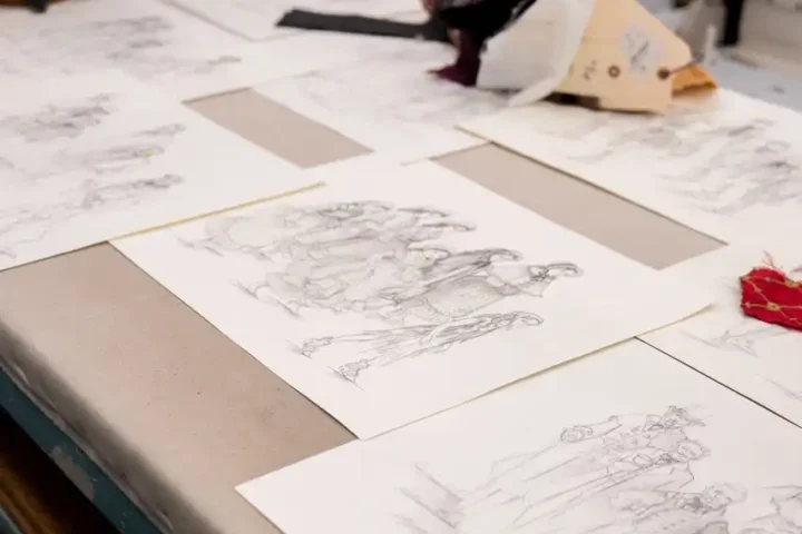 Several detailed pencil sketches of people are laid out on a worktable in a studio setting, surrounded by art supplies and fabric materials, capturing the quiet creativity that reminds us every Christmas we are different.