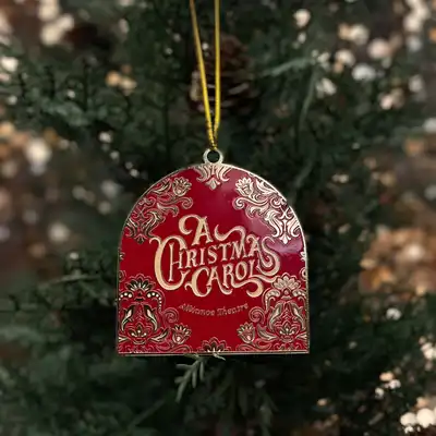 A red and gold Christmas tree ornament with ornate designs and the words "A Christmas Carol" hanging from a yellow string.