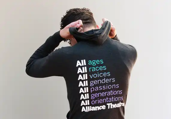 Person facing away, pulling up hood on a sweatshirt that lists "All ages, races, voices, genders, passions, generations, orientations" with "Alliance Theatre" at the bottom.