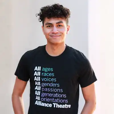 A person stands facing the camera, wearing a black "Alliance Theatre" shirt listing inclusive words such as ages, races, voices, genders, passions, generations, and orientations.