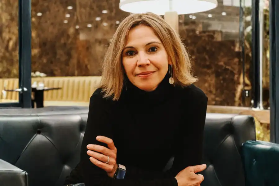 A woman with shoulder-length blonde hair, wearing a black turtleneck and earrings, sits on a dark leather couch with her arms folded, looking at the camera.