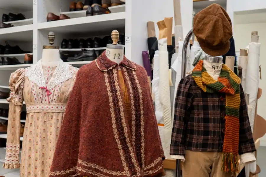 Three mannequins display costumes used in A Christmas Carol—a floral dress, maroon coat, and plaid outfit with a scarf and cap—set before fabric rolls and shelves of shoes.