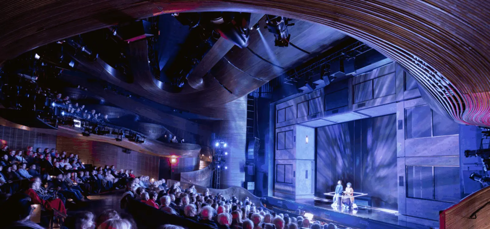 Audience seated in a modern theater watching a stage performance under dramatic blue and purple lighting.