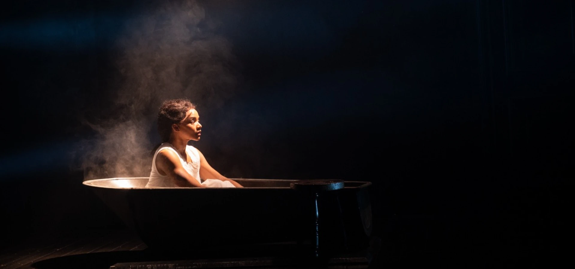 A person sits alone in a metal bathtub filled with steam, illuminated by a spotlight against a dark background, as if caught in the quiet intensity of a Covenant Stream.