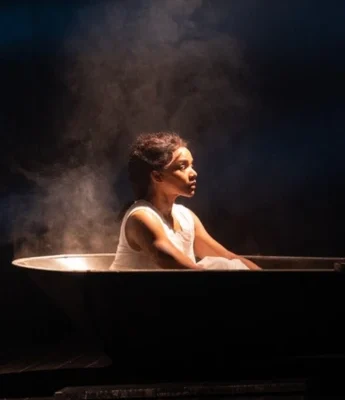 A woman sits in a metal tub with mist rising around her, illuminated by a focused light in the darkness, as if caught in the ethereal flow of a Covenant Stream.