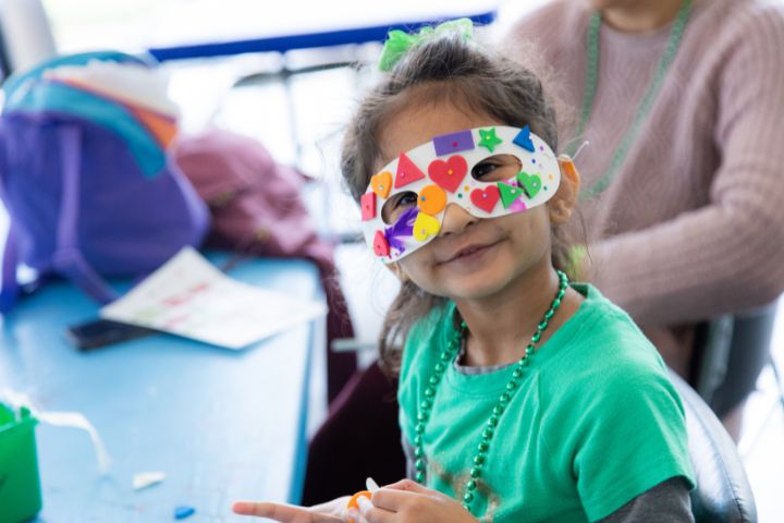 A young girl at the Toddler Takeover Art Festival wears a decorated paper mask and a green shirt, smiling as she sits at a blue table with craft supplies.