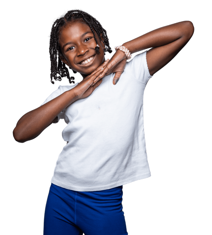Smiling girl with braided hair poses with her hands under her chin, wearing a white t-shirt and blue pants against a plain background.